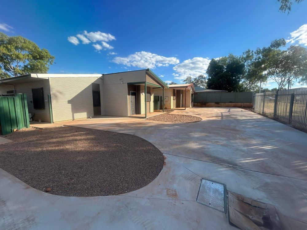 A spacious backyard with a modern single-story house, featuring a large paved area with circular gravel patches. The sky is clear and blue, with a few scattered clouds and trees surrounding the area.