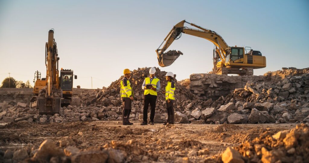 Four construction workers, part of a family-owned business, wear yellow safety vests and helmets as they stand on a rocky site with two large excavators in the background. The sunset casts a warm glow over the machinery and rubble.