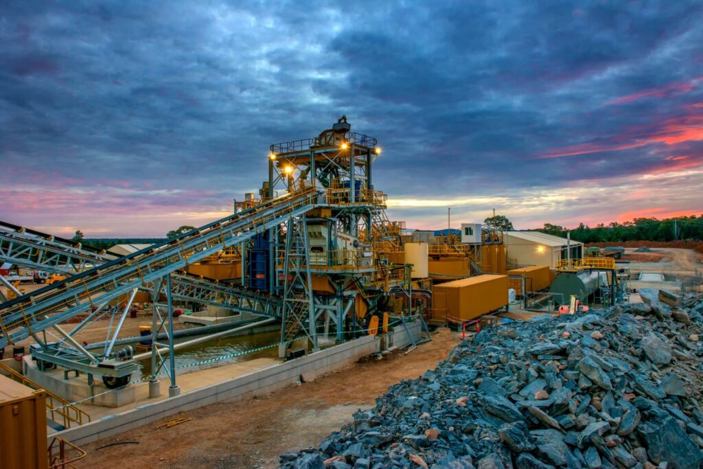 A large industrial mining facility at dusk, rooted in a family-owned business, showcases towering machinery and conveyor belts. Piles of rocks lie in the foreground as a dramatic, cloudy sunset sky paints the backdrop.