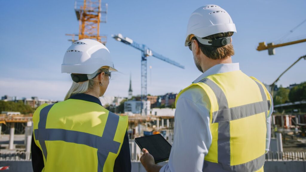 Two construction workers wearing hard hats and reflective vests stand at a family-owned business construction site. One holds a tablet, essential for the day's tasks. Cranes and scaffolding rise in the background under a clear blue sky, showcasing the dedication and pride in their work.
