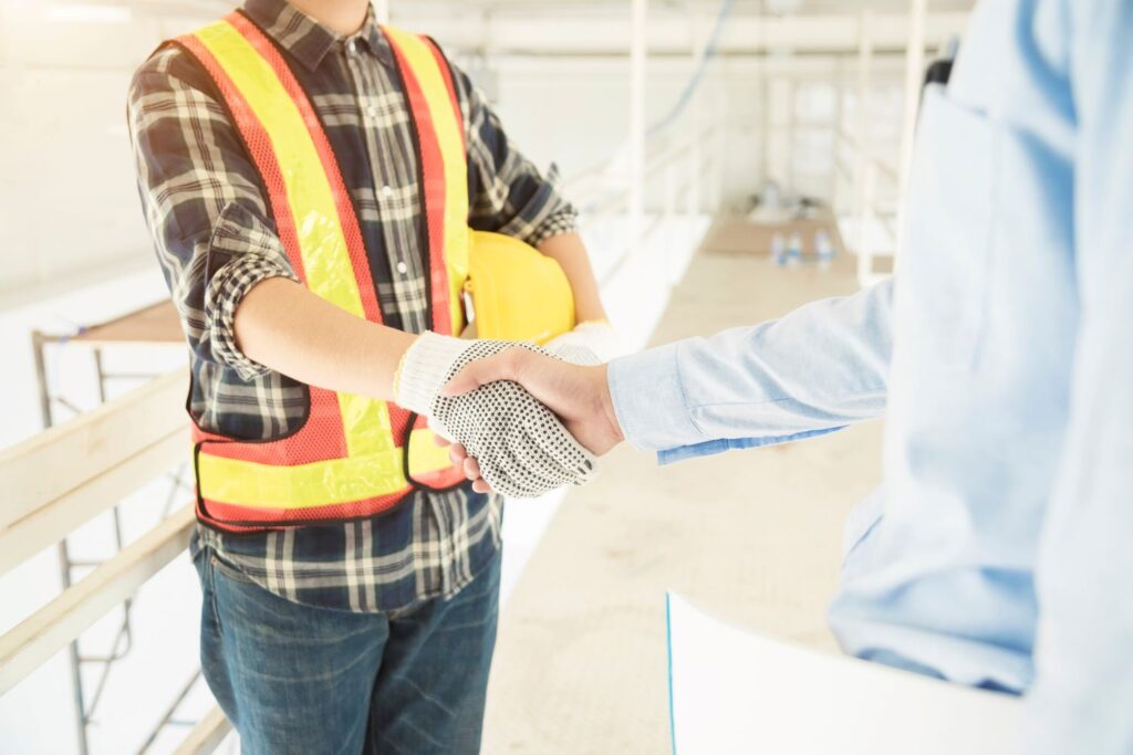 A construction worker in a high-visibility vest and hard hat, representing a family-owned business, shakes hands with a person in a blue shirt on the construction site. Their handshake focuses on symbolizing agreement or collaboration.