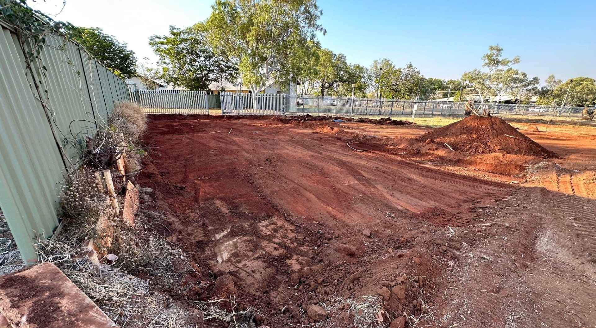 An empty lot with freshly leveled red earth, surrounded by a metal fence and sparse trees. A pile of dirt is visible on the right, and the sky is clear.