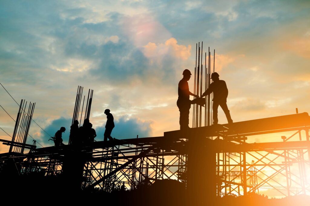 Silhouettes of construction workers on a scaffolding at sunset, with one worker shaking hands with another. The orange sky and clouds create a dramatic backdrop.