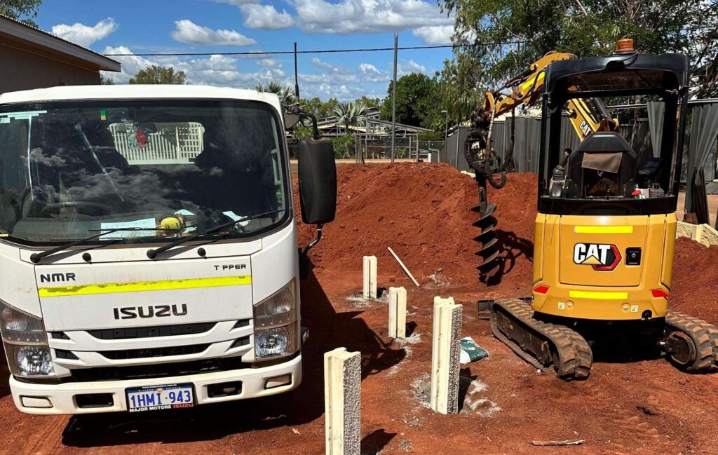 A construction site features a white Isuzu truck parked next to a yellow CAT excavator equipped with an auger, busy with earthworks. The ground is layered in reddish soil, dotted with concrete posts. Trees and a building serve as a backdrop to the industrious scene.