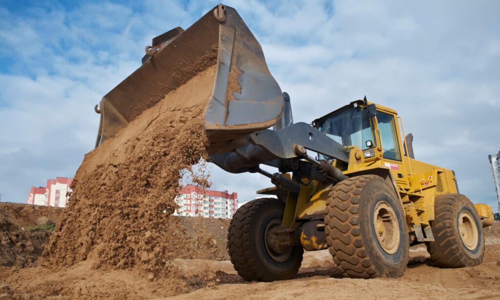 A yellow front loader carries out earthworks, dumping a load of sand at a bustling construction site, with residential buildings standing in the background under a blue, cloudy sky.