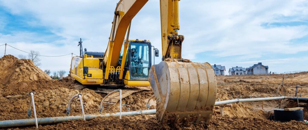 A yellow excavator is busy with earthworks at the construction site, digging into a dirt mound. Piles of earth and metal pipes fill the foreground, while partially constructed buildings rise in the background under the bright blue sky.