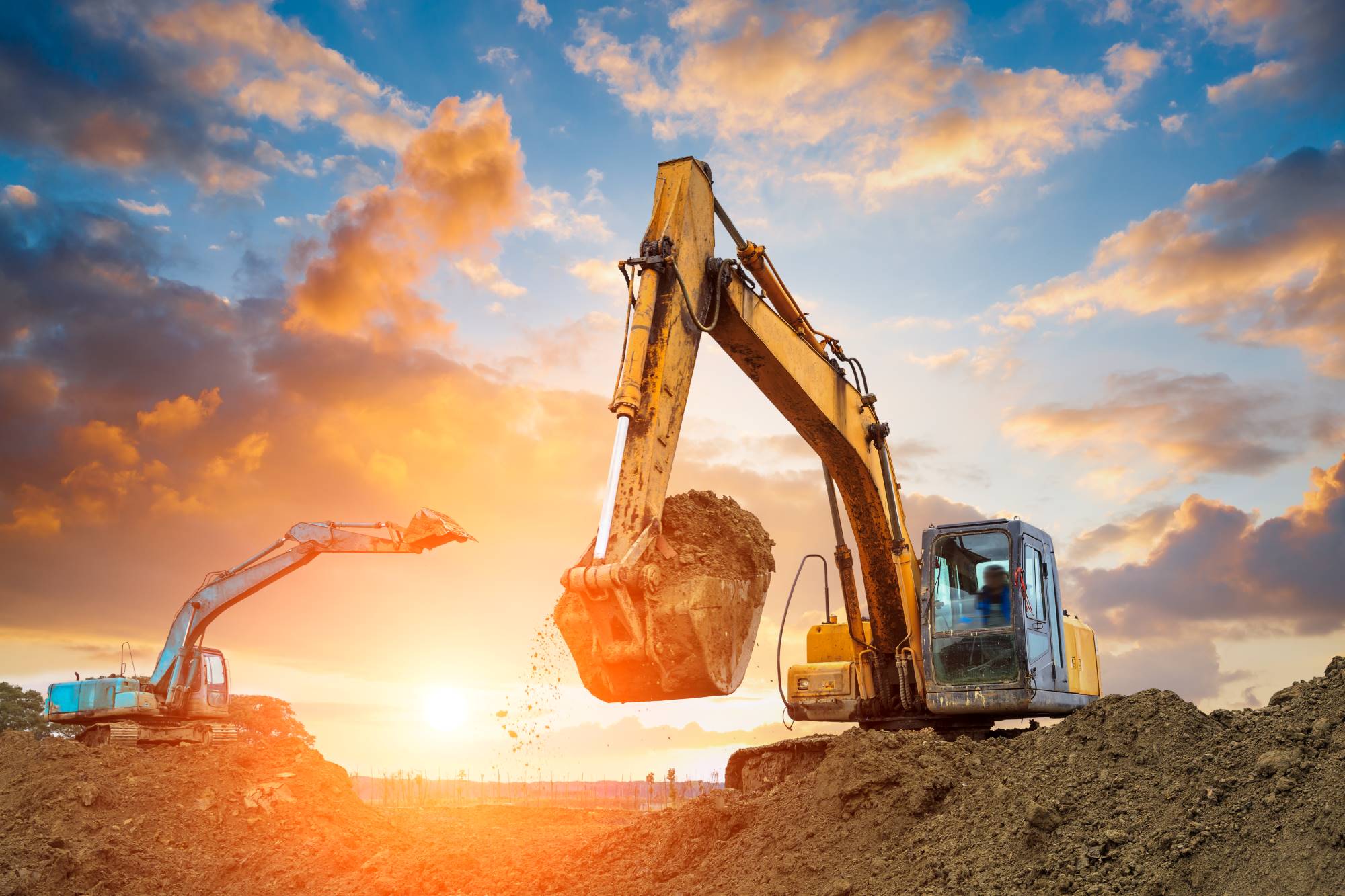 Two excavators in action at a construction site during sunset, with one digging into a pile of dirt while the other stands idle. The vibrant sky, glowing with the colors of the setting sun, forms a breathtaking backdrop. For your construction needs, contact Webley Services today.
