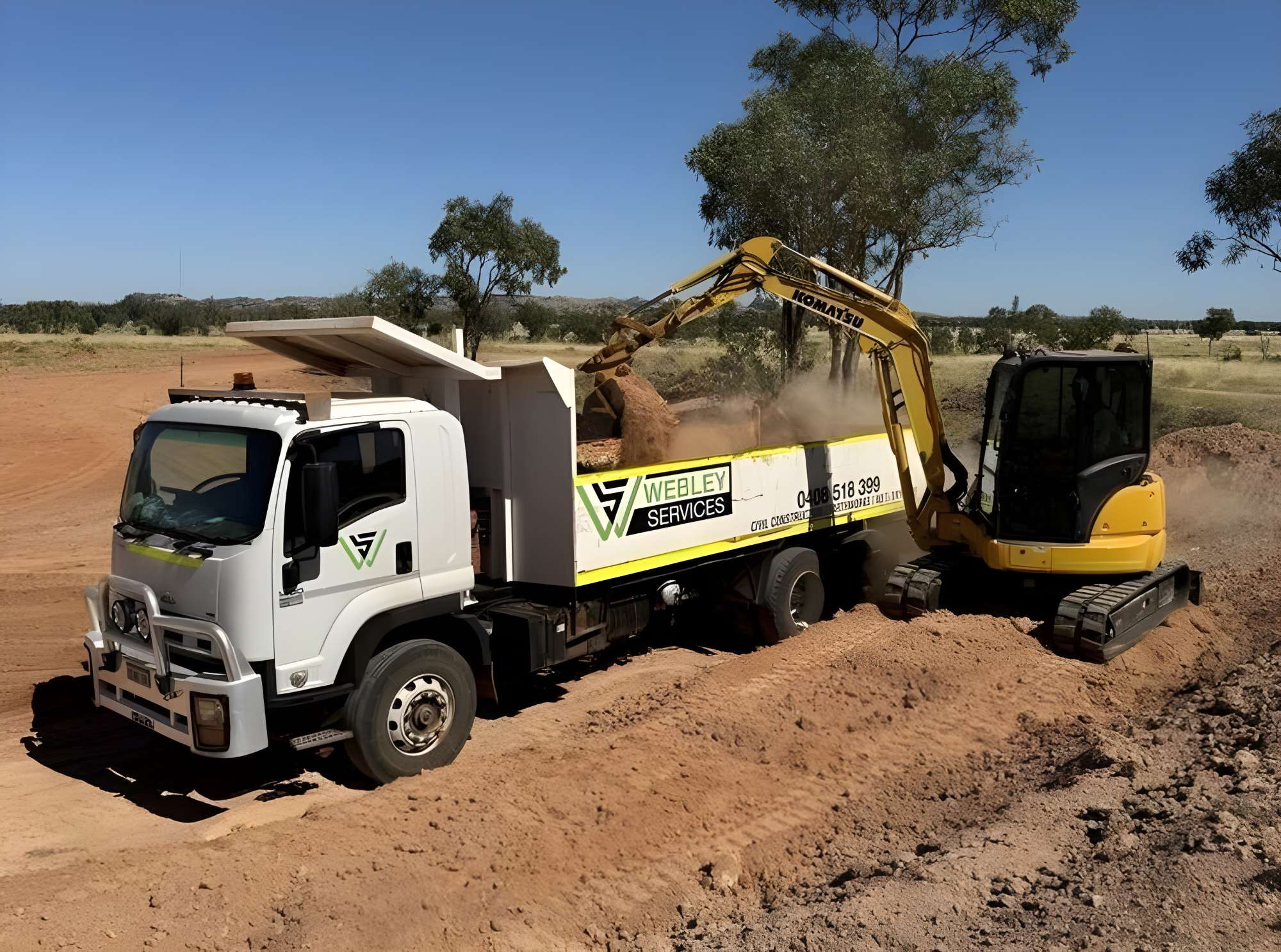 A yellow excavator skillfully performs earthmoving tasks, loading dirt into a white dump truck with "Webley Services" signage. The scene, set on a construction site amid a clear sky and scattered trees, showcases the efficiency of modern haulage operations.