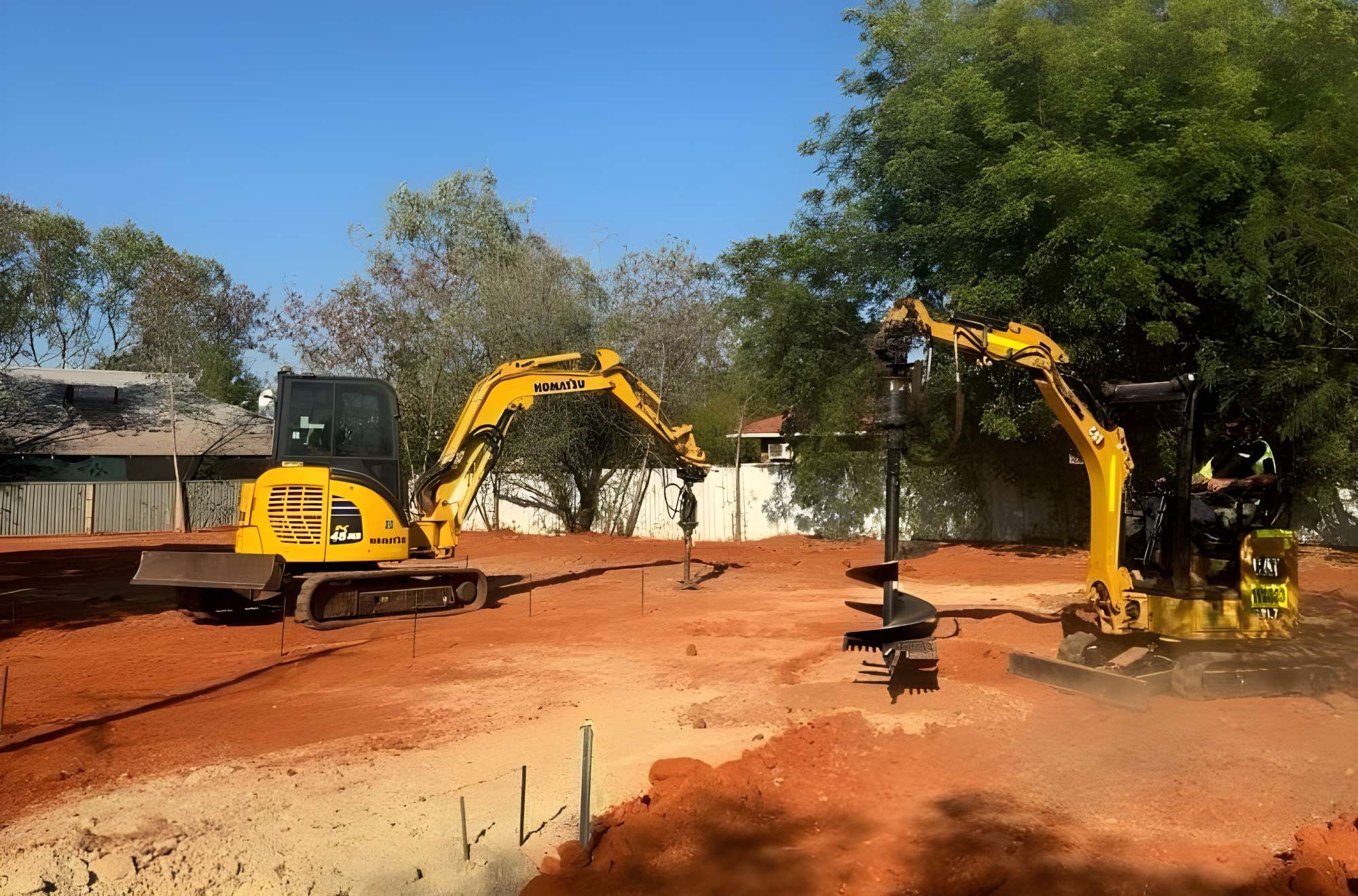 Two yellow excavators operate on a red dirt construction site, engaged in vital earthworks. One is digging while the other moves earth, contributing to the building process. Trees and a fence are visible in the background under a clear blue sky.