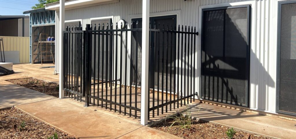 A small fenced area with black metal bars is attached to the side of a building. The structure, hinting at earthworks, has light-colored walls and several windows with dark screens. The concrete ground is bordered by dirt and sparse plants.