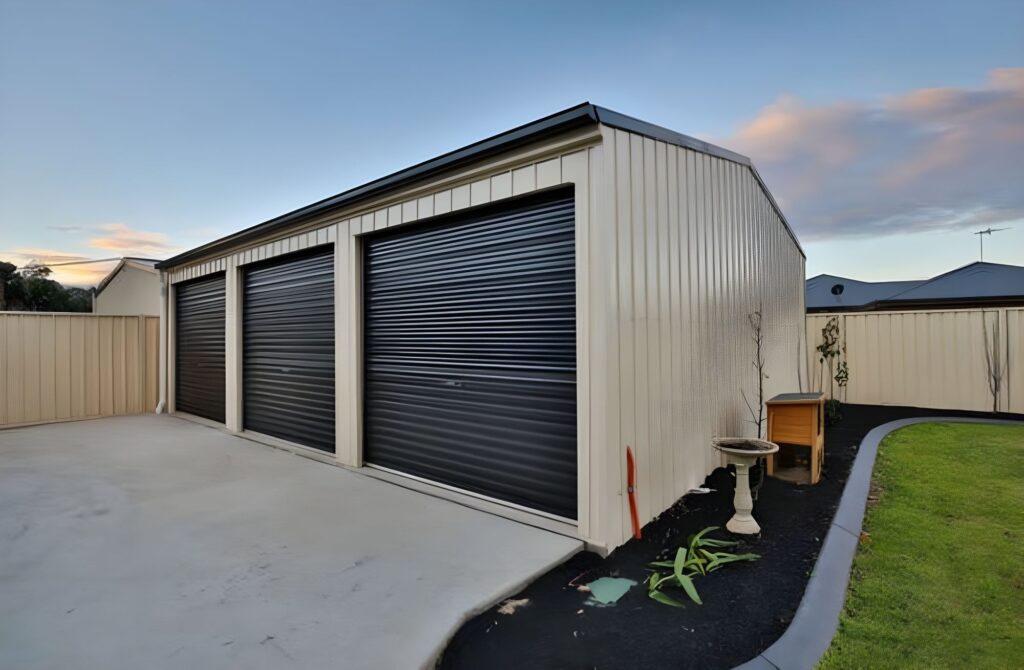 A beige metal garage, a testament to precise building skills, boasts three black roller doors against a clear sky. Nestled on a concrete pad, it's framed by neatly maintained earthworks—a lush lawn and a small garden with a plant and birdbath enhancing its charm.