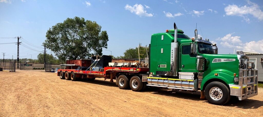 A large green semi-truck with a flatbed trailer is parked on a dirt lot under a clear blue sky. The trailer, partially loaded with various equipment for earthworks, stands ready. Power lines and trees line the background, hinting at nearby building endeavors.